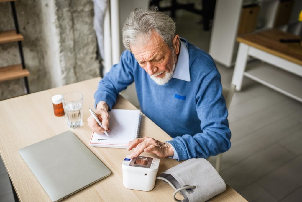 A senior citizen tracking his daily cardiovascular health with a digital upper arm blood pressure cuff and a tracking log.